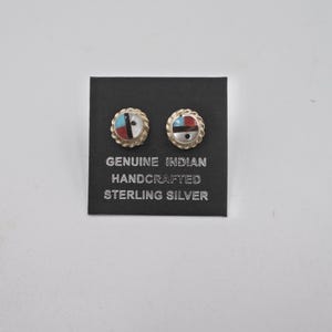 May include: A pair of sterling silver earrings with a Native American design. The earrings are round and have a white, black, red, and blue inlay. The earrings are on a black card that says "Genuine Indian Handcrafted Sterling Silver".
