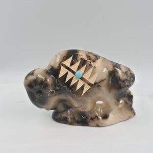 May include: A ceramic sculpture of a bison with a carved geometric design and a turquoise stone accent. The bison is painted in shades of brown and white.