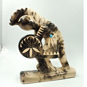 May include: A carved stone sculpture of a Native American figure with a headdress, holding a circular object. The sculpture is beige with black veining and a small turquoise accent. The figure is standing on a rectangular base.