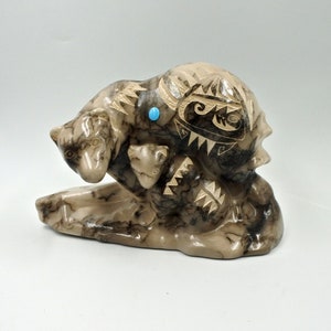 May include: A carved stone sculpture of a mother bear with two cubs. The sculpture is made of a light brown stone with black and white markings. The bear's eyes are inlaid with turquoise stones.