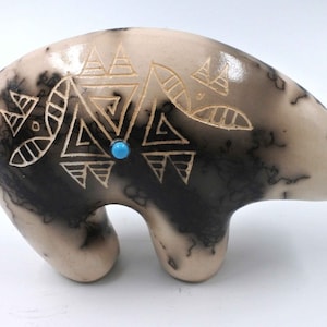 May include: A small ceramic bear figurine with a black and white geometric design and a turquoise stone accent. The bear is standing on all fours and has a smooth, white finish.