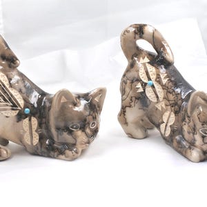 May include: Two ceramic cat figurines featuring a smooth, glossy finish with a marbled black and tan smoke-fired pattern. Each cat is decorated with etched geometric shapes and a small, circular turquoise-colored inlay on the side.