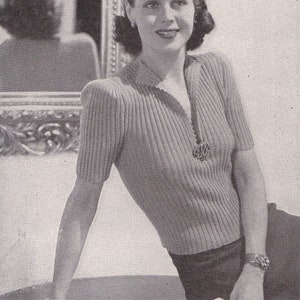 May include: A vintage black and white image of a woman wearing a short-sleeved, ribbed knit top with a decorative pendant. The top has a V-neck and the woman is wearing a bracelet. The woman is smiling and leaning against a surface.