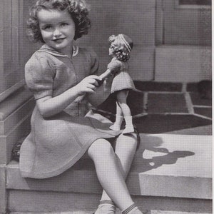 Op de afbeelding: Een jong meisje in een gebreide jurk zit op een trede en houdt een pop vast die een vergelijkbare jurk draagt. De pop heeft een witte matrozenkraag en het meisje draagt zwarte schoenen met witte bandjes.