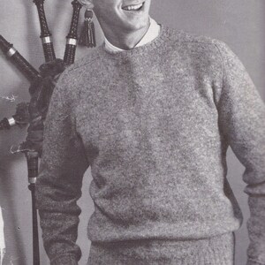 May include: A man wearing a grey sweater with a crew neck and a white undershirt. He is standing next to a set of bagpipes.