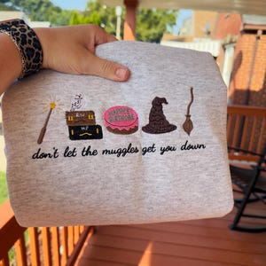 May include: A light gray sweatshirt featuring embroidered icons including a wand, a white owl on two stacked trunks, a pink cake with the text HAPPEE BIRTHDAE, a brown wizard hat, and a broomstick, with the text don't let the muggles get you down.