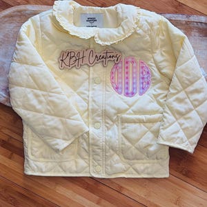 May include: A pale yellow quilted jacket featuring a ruffled collar, long sleeves, and two front pockets. The jacket has a snap-button closure and a pink and white gingham monogram patch on the chest. A wooden sign reads KBH Creations.