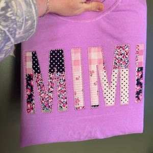 May include: A lilac-colored sweatshirt with the word "MIMI" appliquéd across the front. The letters are made from various patterned fabrics, including floral, polka dot, and gingham designs. The sweatshirt is folded.