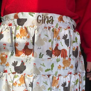 May include: A white apron with a repeating pattern of chickens, roosters, and chicks in various colors. The apron has three ruffled tiers and the name "Gina" embroidered in brown thread across the top.