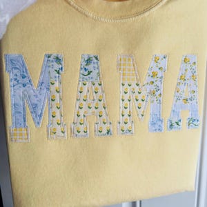 May include: A light yellow sweatshirt with the word "MAMA" appliqued across the front. The letters are made of fabric with floral and gingham patterns in blue and yellow. The sweatshirt has a crew neck and long sleeves.