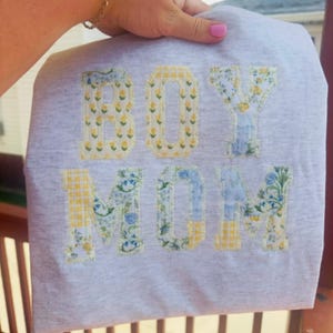 May include: A light gray t-shirt featuring the text BOY MOM appliqued in block letters. The letters are crafted from fabric with yellow and blue floral and gingham patterns, finished with a visible stitched border around each letter.