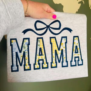 Op de afbeelding: Een lichtgrijs sweatshirt met het woord "MAMA" geborduurd in marineblauw met bloemen- en geel geruite patronen. Boven het woord is een marineblauwe strik geborduurd. Het sweatshirt wordt door een persoon vastgehouden.