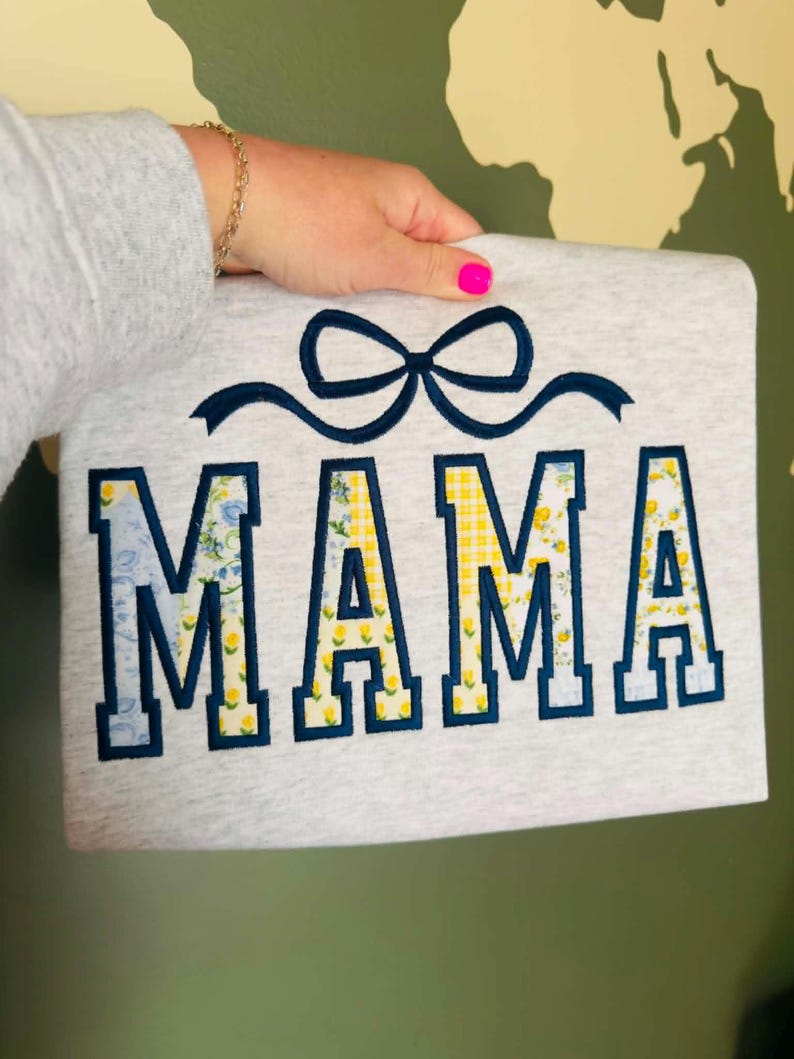 Op de afbeelding: Een lichtgrijs sweatshirt met het woord "MAMA" geborduurd in marineblauw met bloemen- en geel geruite patronen. Boven het woord is een marineblauwe strik geborduurd. Het sweatshirt wordt door een persoon vastgehouden.