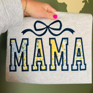 Op de afbeelding: Grijze sweatshirt met het woord "MAMA" in marineblauwe blokletters. De letters zijn gevuld met bloemen- en geel geruite patronen. Boven het woord is een marineblauwe strik geborduurd.