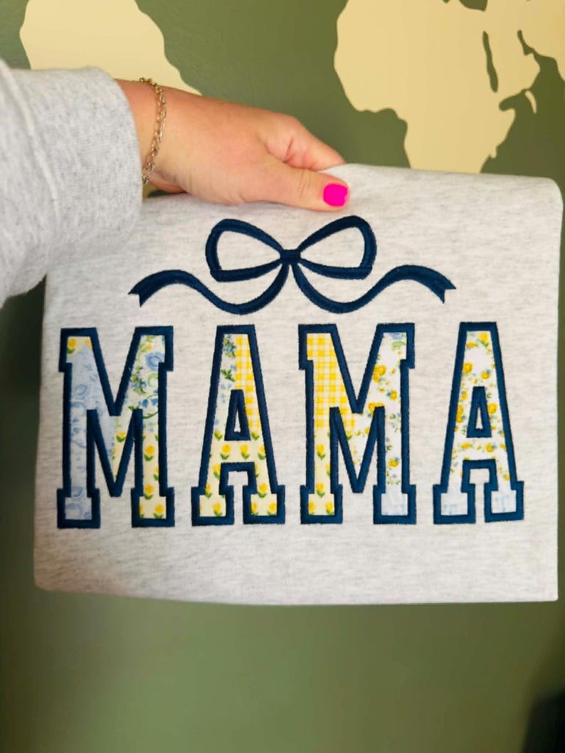 Op de afbeelding: Grijze sweatshirt met het woord "MAMA" in marineblauwe blokletters. De letters zijn gevuld met bloemen- en geel geruite patronen. Boven het woord is een marineblauwe strik geborduurd.