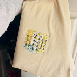 May include: Cream-colored tote bag with a decorative patch. The patch features the embroidered initials "KHH" in a blue script font, set against a background of yellow gingham and floral patterns. The bag is held up by a person.