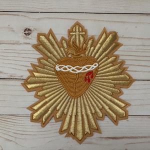 Puede incluir: Parche bordado con un coraz&oacute;n dorado con una corona de espinas blanca y gotas de sangre rojas. El coraz&oacute;n est&aacute; rodeado de un sol radiante dorado.