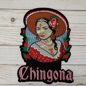 May include: Embroidered iron-on patch featuring a woman wearing a red dress and a large brown hat with a red flower. The woman has a red flower in her hair and a red dot on her forehead. The patch has the word "Chingona" in pink lettering.