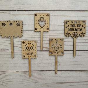 Peut inclure: Cinq pancartes en bois découpées avec différents motifs, dont un cœur, un crâne, une guitare et une bannière qui dit "Dia de Muertos". Les pancartes sont sur des bâtonnets en bois et sont parfaites pour décorer une fête ou un événement.