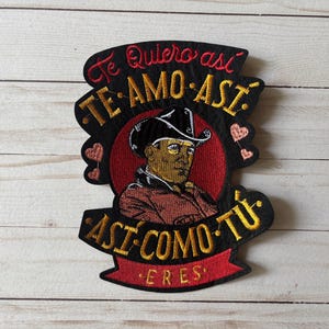 May include: Embroidered patch featuring a man in a cowboy hat, with Spanish text. The black patch has gold and red detailing, including the phrases "Te Quiero así" and "Te Amo Así". The design includes heart motifs.