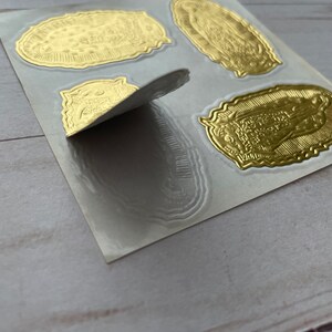 May include: A sheet of gold foil stickers featuring an image of the Virgin Mary. The stickers are on a white backing sheet and are ready to be peeled off and used.
