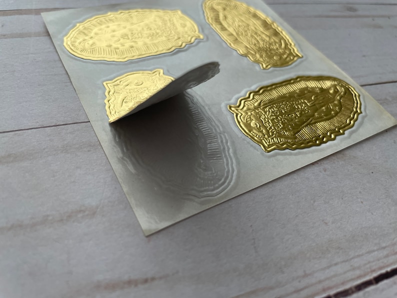 May include: A sheet of gold foil stickers featuring an image of the Virgin Mary. The stickers are on a white backing sheet and are ready to be peeled off and used.