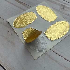 May include: A sheet of gold foil stickers featuring an image of the Virgin Mary. The stickers are oval shaped and have a raised, textured design.
