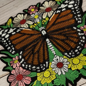 May include: Embroidered butterfly patch with brown wings, white and black accents, and a border of white sequins. The butterfly is surrounded by colorful embroidered flowers in pink, yellow, and white.