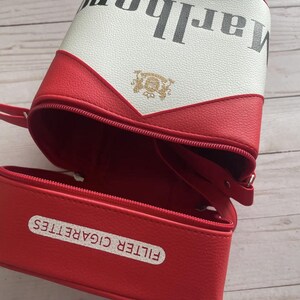 May include: A red and white cigarette-shaped bag with a zipper closure. The bag has the word "Marlboro" printed on it in black. The bottom of the bag reads "FILTER CIGARETTES".
