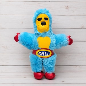 Peut inclure: Une peluche en forme de catcheur avec une fourrure bleu vif, un masque jaune avec des ouvertures noires pour les yeux et un empiècement jaune sur la poitrine. Elle possède des gants et des bottes rouges métallisés, ainsi qu'une ceinture rouge.
