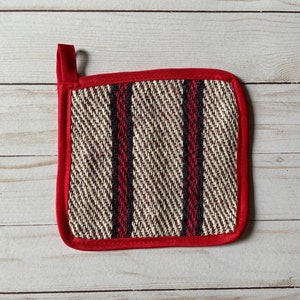 Puede incluir: Un agarrador de olla a rayas rojas y blancas con un lazo rojo para colgar. El agarrador de olla está hecho de tejido tejido y tiene un borde rojo.