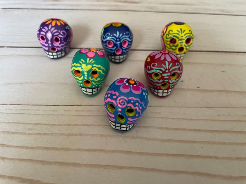 May include: Six brightly coloured ceramic sugar skulls, each hand-painted with unique designs. The skulls feature a variety of colours including purple, blue, green, yellow, and red. They have white teeth and are decorated with floral and geometric patterns.