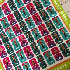 May include: A white square scarf with a repeating pattern of FIFA World Cup trophy graphics in teal, pink, and purple. The scarf has green and orange borders with text that reads "Guadalajara - Monterrey - CDMX - Guadalajara."