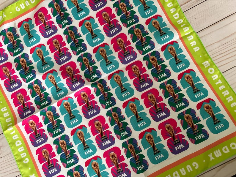 May include: A white square scarf with a repeating pattern of FIFA World Cup trophy graphics in teal, pink, and purple. The scarf has green and orange borders with text that reads "Guadalajara - Monterrey - CDMX - Guadalajara."