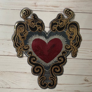 May include: A heart-shaped applique with a red beaded heart center, surrounded by silver, gray, and black sequins. Gold sequin embellishments and black mesh outline the design. Decorative craft item.