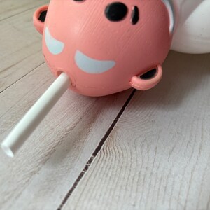 Peut inclure: Un personnage rose et blanc avec une paille blanche qui sort de sa bouche. Le personnage est allong&eacute; sur une surface en bois clair.