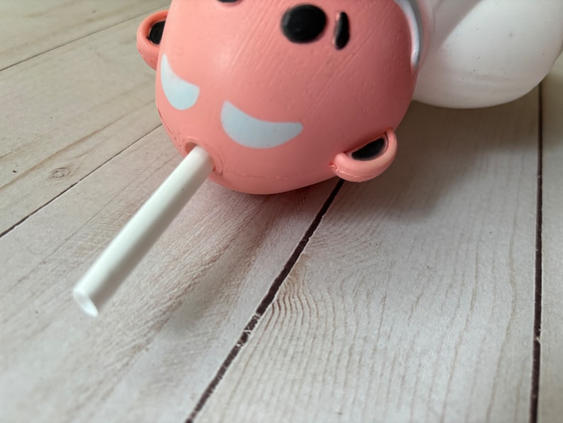 Peut inclure: Un personnage rose et blanc avec une paille blanche qui sort de sa bouche. Le personnage est allong&eacute; sur une surface en bois clair.