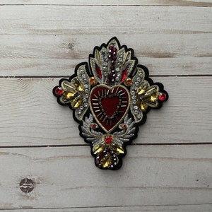 May include: A red heart with gold and silver embellishments, surrounded by a black fabric with gold and silver embroidery. The heart is surrounded by a crown of thorns.