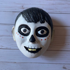 May include: A white mask with black and red painted details depicting a skull face with large eyes. The mask has black hair and a painted smile with yellow teeth.
