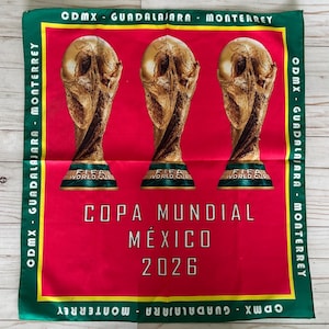 May include: A red and green square textile featuring three gold FIFA World Cup trophies. The text "COPA MUNDIAL M&Eacute;XICO 2026" is centered below the trophies. The border includes the text "CDMX - GUADALAJARA - MONTERREY".