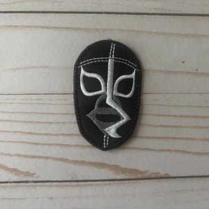 Puede incluir: Parche bordado negro y blanco con un diseño de máscara estilizada. La máscara presenta detalles blancos y grises sobre un fondo oscuro, incluyendo formas de ojos y una línea vertical central. Ideal para ropa.
