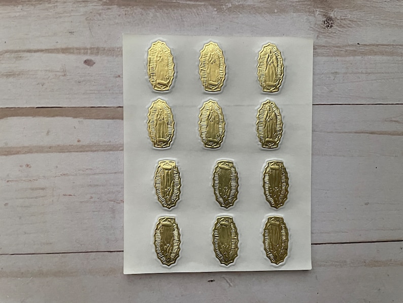 May include: A sheet of gold-coloured stickers featuring an image of the Virgin Mary. The stickers are arranged in three rows of four, with each sticker depicting the Virgin Mary in a similar pose.