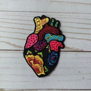Puede incluir: Parche bordado con forma de corazón humano, con contorno negro y relleno de coloridos diseños florales y anatómicos. El parche incluye tonos de rojo, amarillo, rosa, azul y verde.