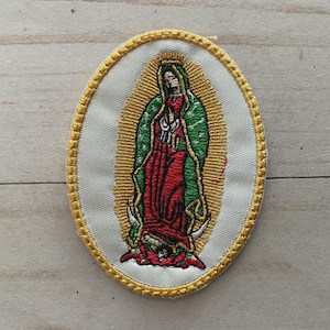 Op de afbeelding: Geborduurde patch met de Maagd van Guadalupe. De ovale patch heeft een witte achtergrond met een gouden rand. De afbeelding toont de Maagd Maria in een rode jurk en groene mantel, omringd door een gouden halo.