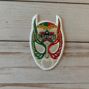 Könnte beinhalten: Ein gestickter Aufnäher in Form einer Luchador-Maske mit cremefarbenem, grünem und rotem Garn. Das Design zeigt einen zentralen Stern, den Schriftzug EL GRANDE in schwarzen Buchstaben und symmetrische flügelartige Muster mit weißem Rand.