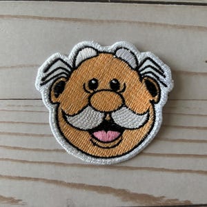 May include: Embroidered patch featuring a cartoon face with a tan complexion, black eyes, and a white mustache. The patch has a white border and a pink tongue. The design is stitched with black, white, tan, and pink thread.