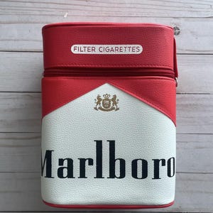 May include: A red, white, and black cigarette case with the word "Marlboro" in black lettering. The case has a zipper closure and the words "FILTER CIGARETTES" are printed on the top.