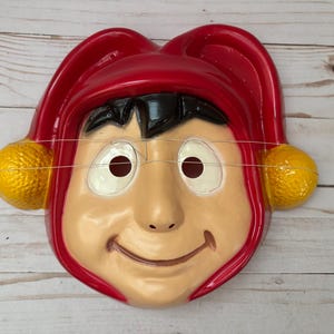 Puede incluir: Una máscara de bufón de plástico roja y amarilla con una cara sonriente y cabello negro. La máscara tiene agujeros para los ojos y una cuerda atada a los lados.