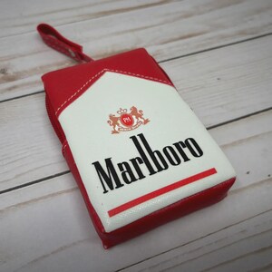 May include: A red and white zippered pouch with a design resembling a Marlboro cigarette pack. The pouch features the Marlboro logo in black, a red stripe, and a small handle. The background is a light wood grain.