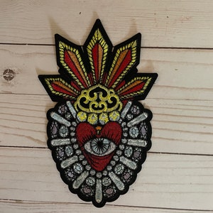 Puede incluir: Parche bordado para planchar con un corazón rojo con un ojo en el centro, rodeado de un diseño de cuentas blancas y plateadas. El corazón está coronado con un diseño de llamas amarillas y naranjas.